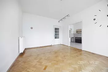 Světlý obývací pokoj s parketovou podlahou, moderní osvětlení a vstup do kuchyně, bílá výmalba pro svěží vzhled.