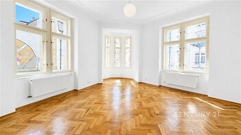 Světlý obývací pokoj s parketovou podlahou, velkými okny a vysokými stropy, vytvářející moderní a vzdušný prostor.