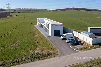 Průmyslová hala s moderním designem, velkým parkovištěm a novou fasádou, obklopená zelení a snadným přístupem po asfaltové cestě.