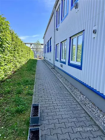 Průmyslový areál s dlážděným chodníkem vedle budovy se světlou fasádou a modrými rámy oken, obklopený zeleným živým plotem.