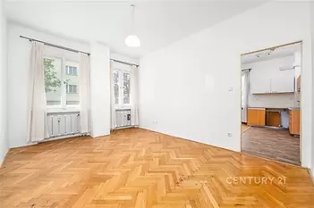 Světlý obývací pokoj s parketovou podlahou a velkými okny přechází do moderní kuchyně.