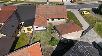 Venkovní prostor s trávníkem, dvěma domy se šikmými střechami a přístřeškem, dlážděná příjezdová zóna, klidná rezidenční lokalita.
