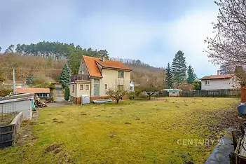 Prostorná zahrada s rodinným domem, doplněná zeleným trávníkem a vzrostlými stromy, nabízí klidné prostředí a soukromí pro bydlení.