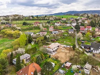 Letecký pohled na vesnickou oblast s rozptýlenými domy a zahradami obklopenými zelení, s výhledem na kopcovitou krajinu v pozadí.